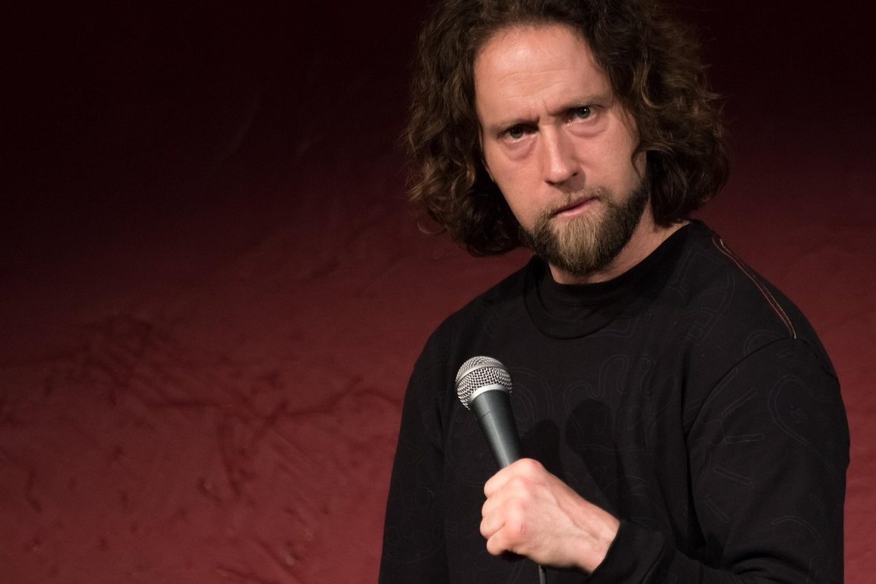A comedian with curly hair and a beard holding a microphone against a red backdrop.