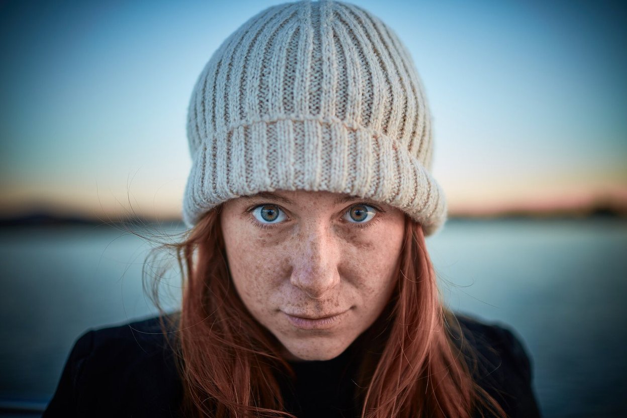 A young woman with red hair and freckles wearing a knit hat by a lake