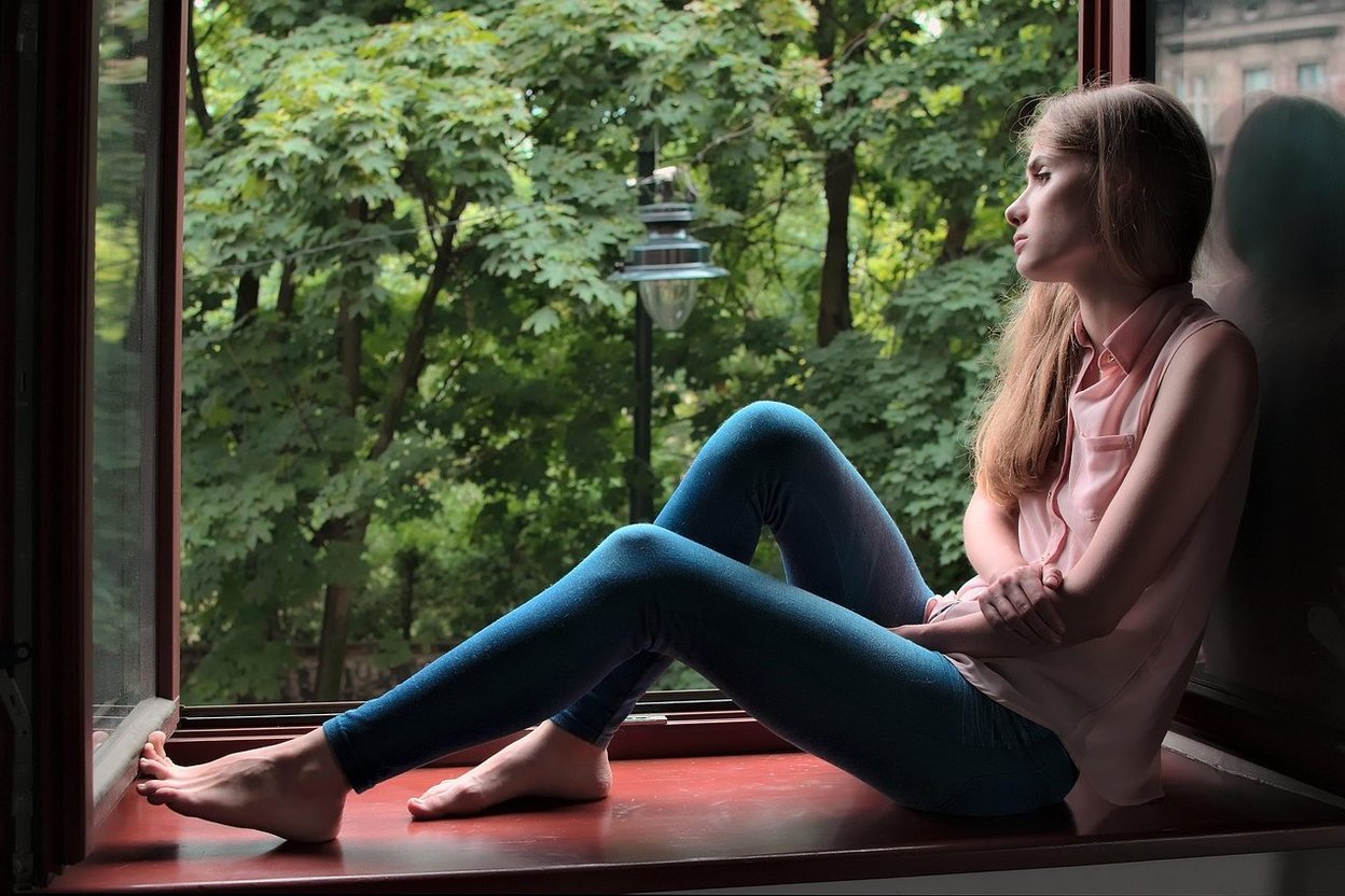 A young woman sitting quietly on a windowsill, looking out at green trees.