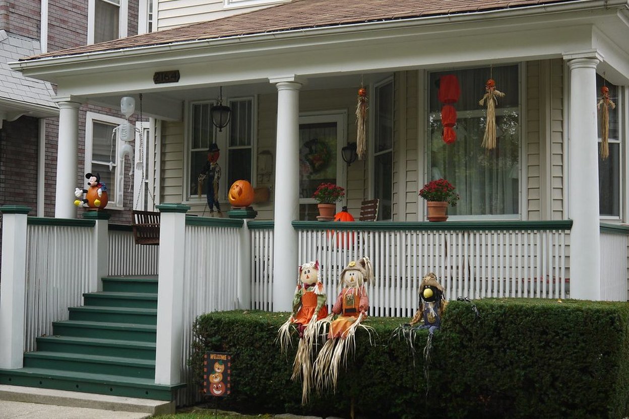 A family in costumes holding trick-or-treat bags on a porch decorated with pumpkins and string lights at dusk.