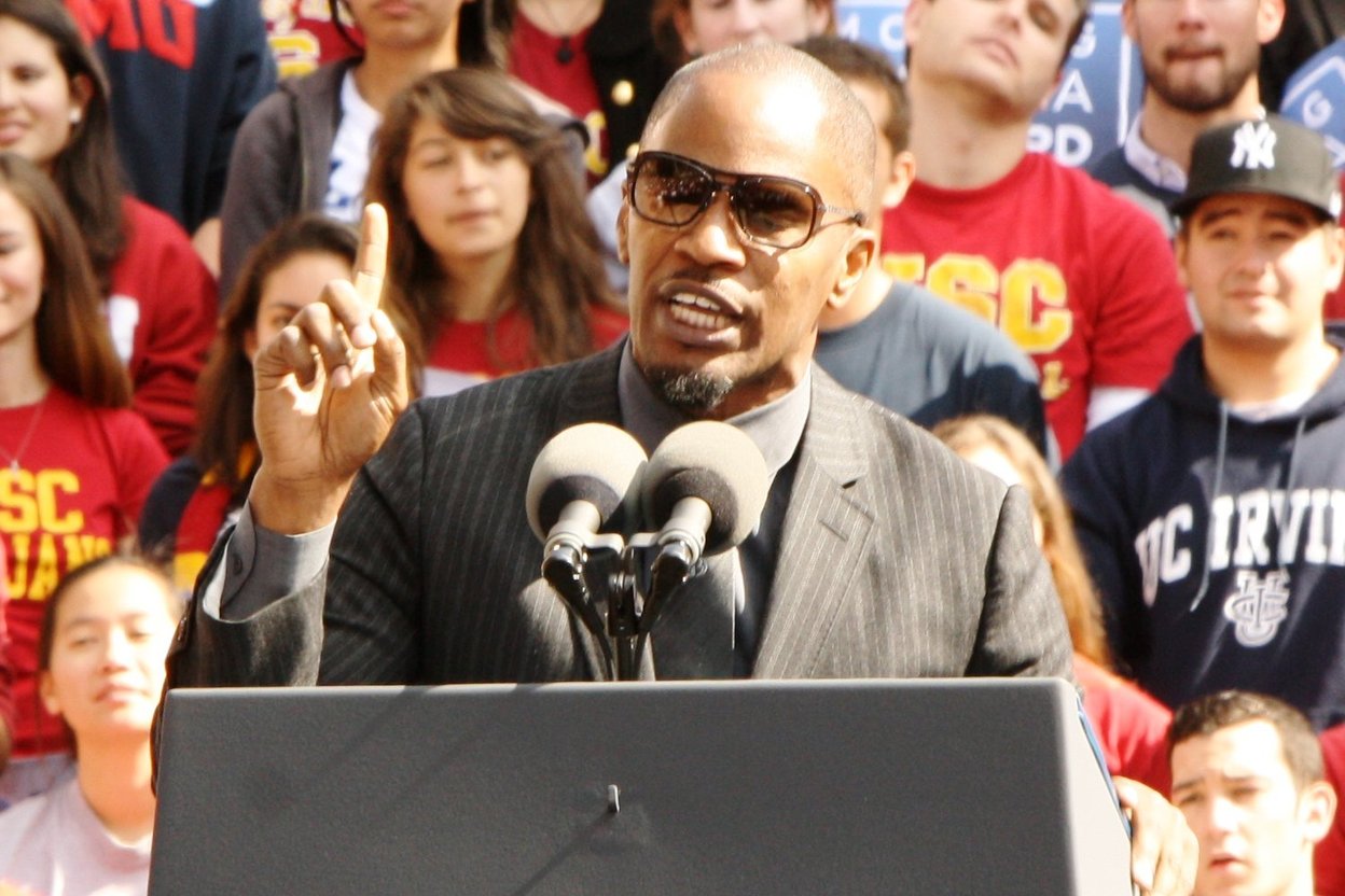 Jamie Foxx in a suit and sunglasses speaking into microphones at a podium in front of a crowd.
