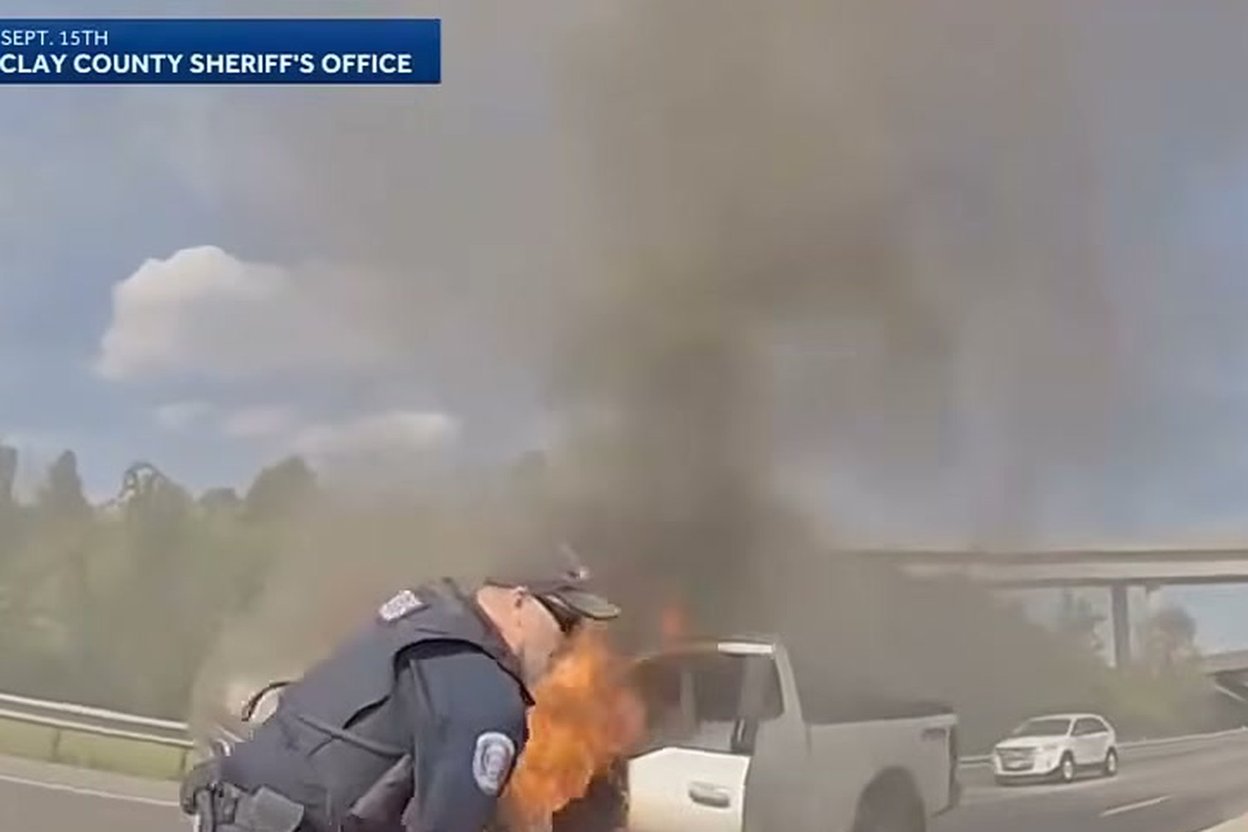 Dashcam view of a police officer pulling a driver away from a burning white pickup on an interstate