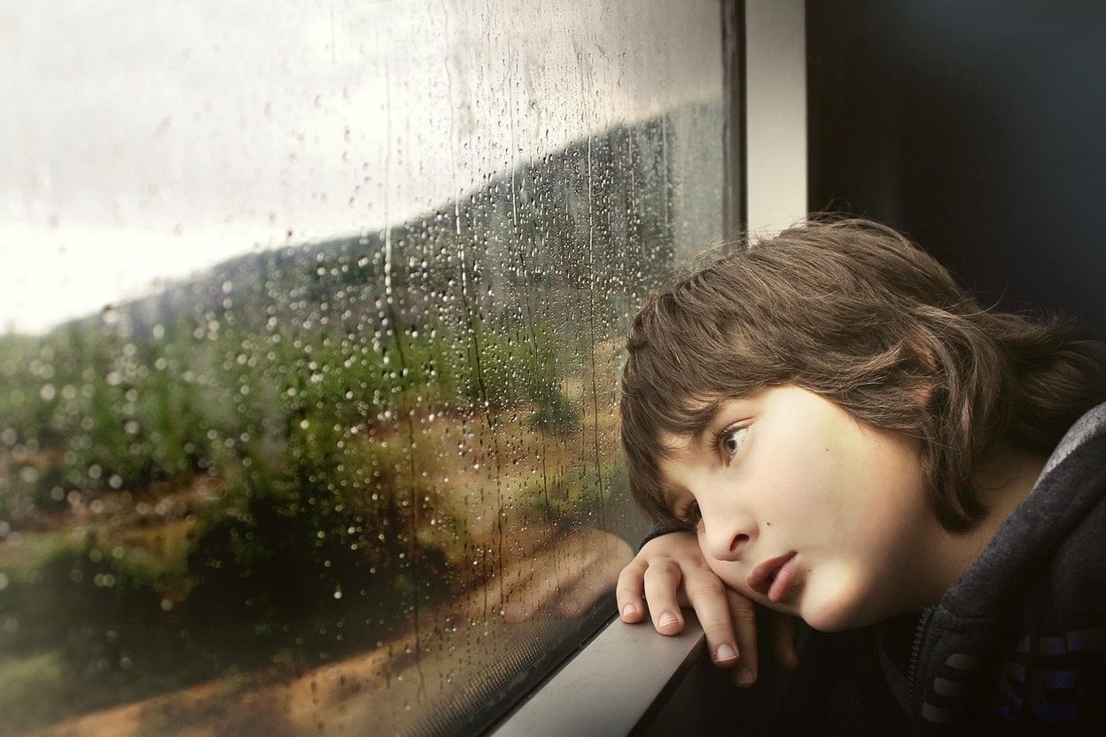 A young person gazes out a rain-splattered window at a blurred landscape.