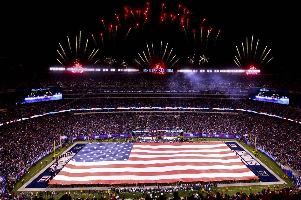 A giant American flag spread across the field of a packed NFL stadium at night, with fireworks overhead.