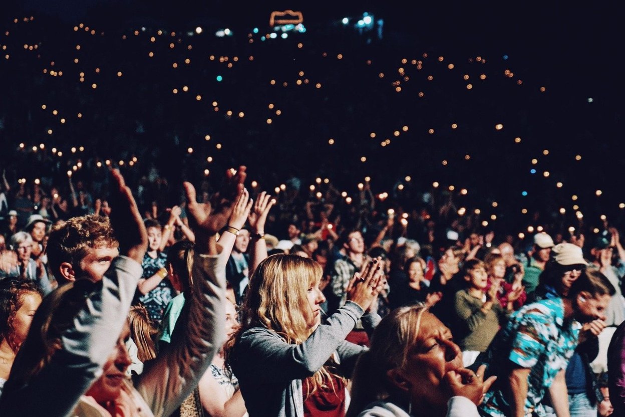 A huge crowd at a nighttime show, many with hands raised under the lights.